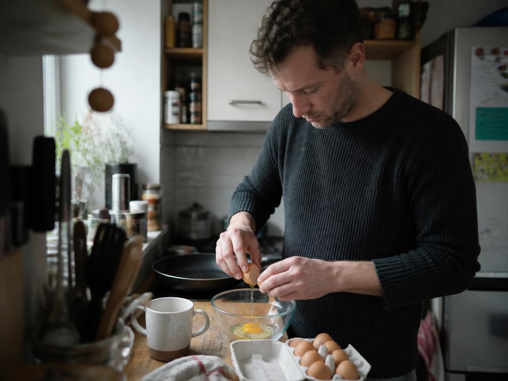 A man cracking a brown egg into a glass bowl in a lived-in home kitchen