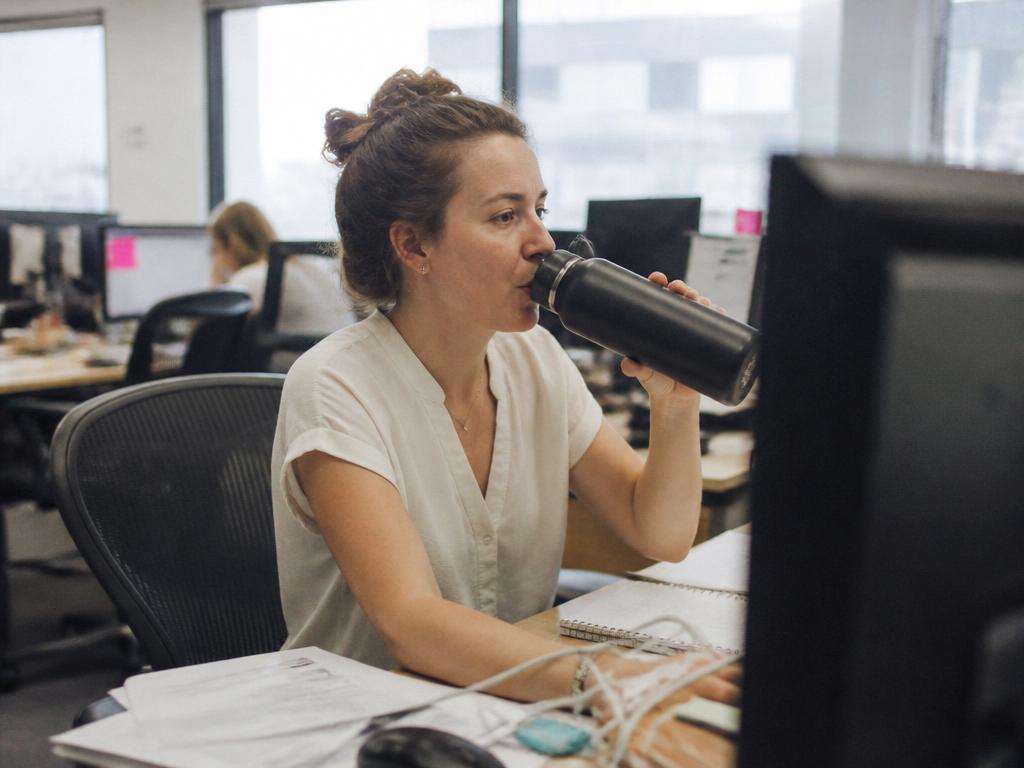 A woman drinking from a large reusable water bottle while working at an office desk