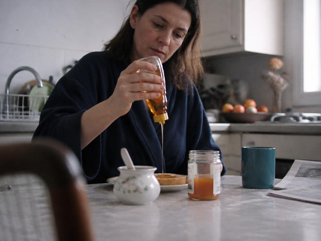 A woman drizzling honey onto toast beside a sugar bowl at a kitchen table